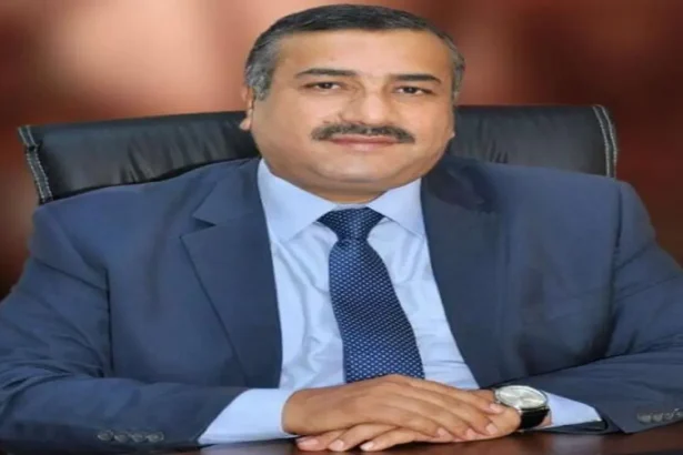 صورة شخصية للأستاذ حاكم المحاميد يرتدي بدلة رسمية مع ربطة عنق ذهبية، متبسماً أمام مبنى سكني في وضح النهار.