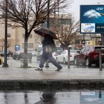 تأثر الأردن بالمنخفض الجوي مع عبور مشاة يحمل مظلة في شارع ممطر بجانب لوحة إعلانية لطقس الأردن