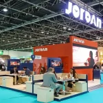 منظر بانورامي لجناح الأردن المزين باللافتات المضيئة في معرض تجاري دولي للسياحة، يظهر زواراً يناقشون ويفاوضون في مناطق الجلوس.