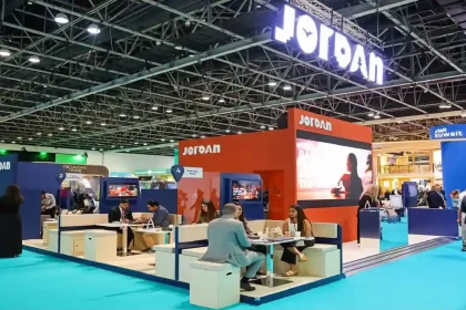 منظر بانورامي لجناح الأردن المزين باللافتات المضيئة في معرض تجاري دولي للسياحة، يظهر زواراً يناقشون ويفاوضون في مناطق الجلوس.