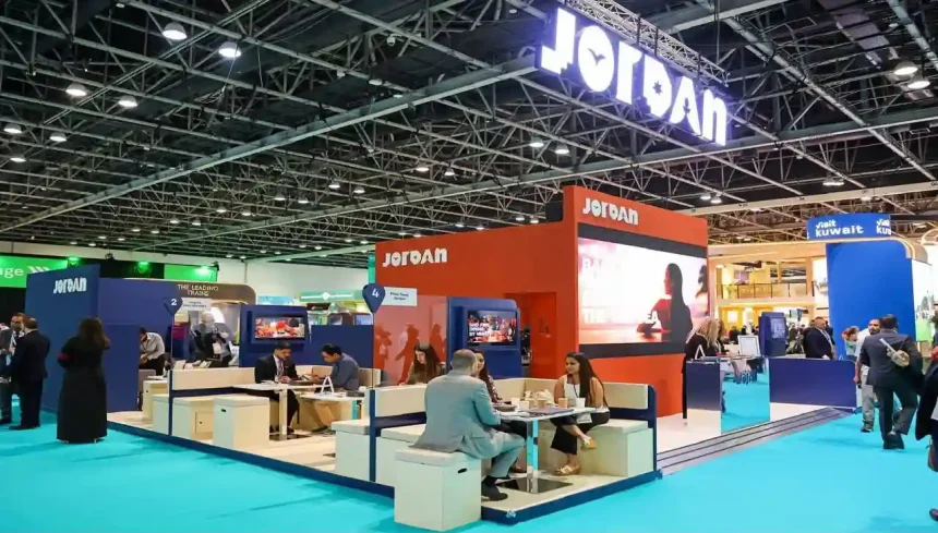 منظر بانورامي لجناح الأردن المزين باللافتات المضيئة في معرض تجاري دولي للسياحة، يظهر زواراً يناقشون ويفاوضون في مناطق الجلوس.