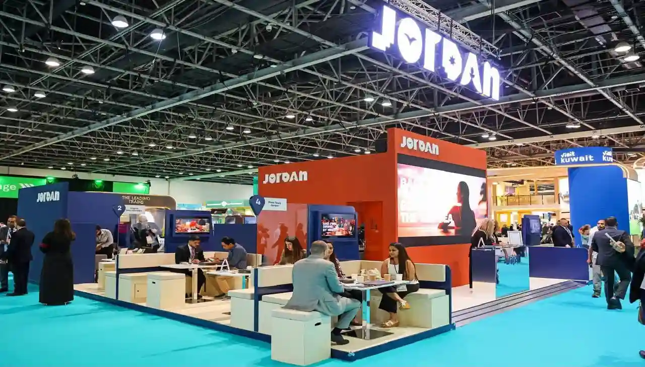 منظر بانورامي لجناح الأردن المزين باللافتات المضيئة في معرض تجاري دولي للسياحة، يظهر زواراً يناقشون ويفاوضون في مناطق الجلوس.