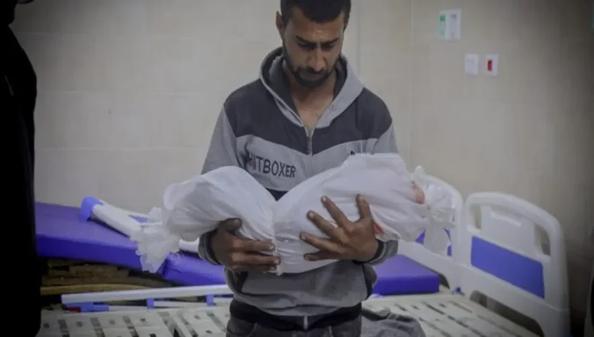 أب فلسطيني يحمل جثمان طفله ملفوفاً بالكفن الأبيض ضمن شهداء غزة بعد الهدنة في مستشفى مكتظ بالجرحى