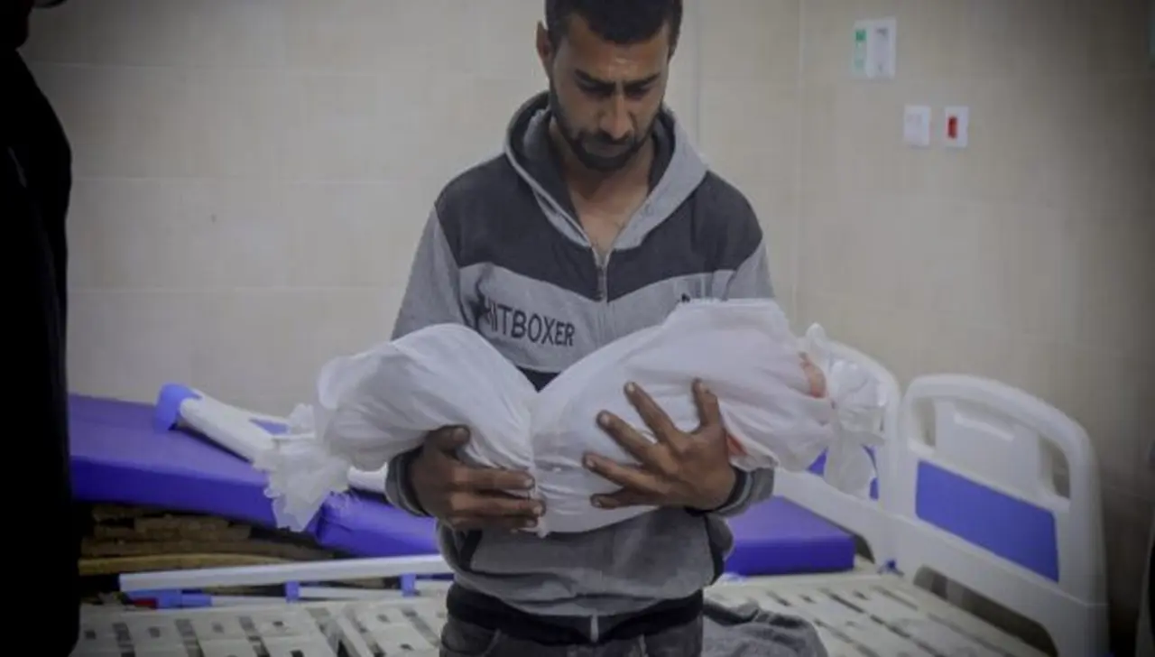 أب فلسطيني يحمل جثمان طفله ملفوفاً بالكفن الأبيض ضمن شهداء غزة بعد الهدنة في مستشفى مكتظ بالجرحى
