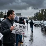 رجل في المقدمة يحمل صحيفة "الطليعة نيوز" وعنوانها "الحالة الجوية وتأثيرها على المملكة" في شارع ممطر.