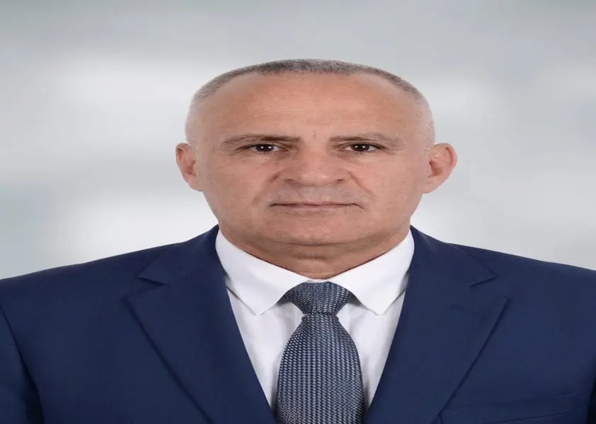 صورة شخصية رسمية ليونس العبادلة يرتدي بدلة كحلية وربطة عنق رمادية خلفية رمادية فاتحة